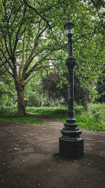 A tall, black, vintage-style lamppost stands near a winding dirt pathway in a lush, green park setting. The lamppost features an ornate, decorative base and a lantern at the top with glass panels. Sur