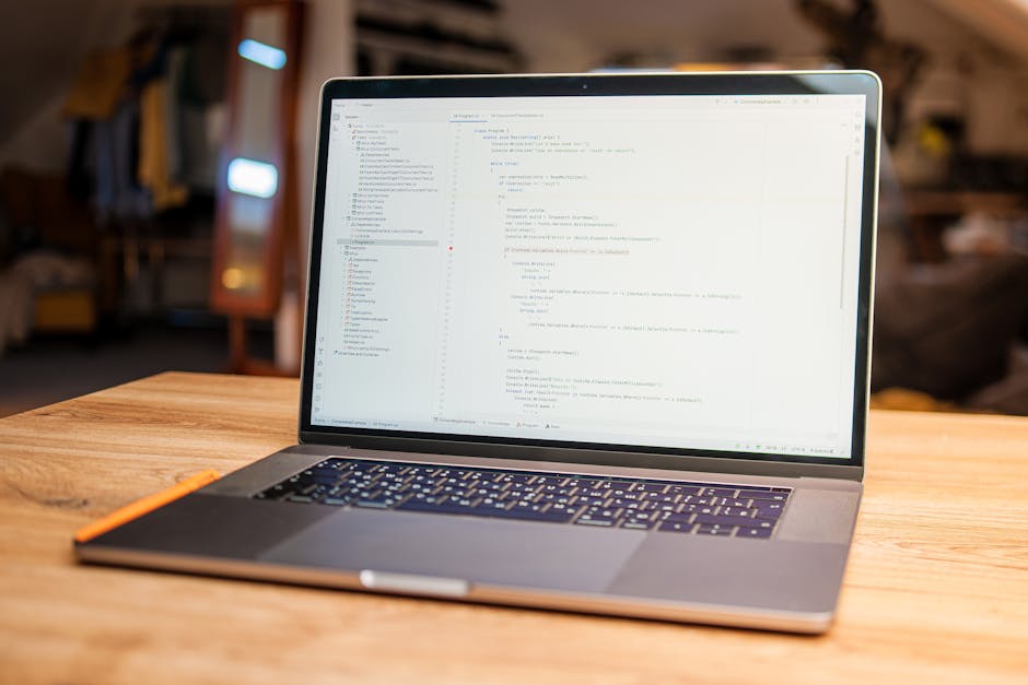 A silver laptop placed on a wooden table displays a screen filled with lines of code, set in a cozy indoor environment with warm ambient lighting. In the background, blurred elements of home decor and